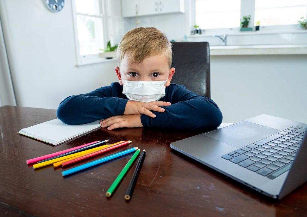 Child wearing mask at table with laptop and colored pencils